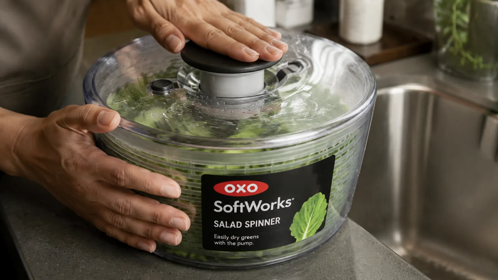 Hands pressing the pump button on an OXO salad spinner lid with wet lettuce visible inside the bowl