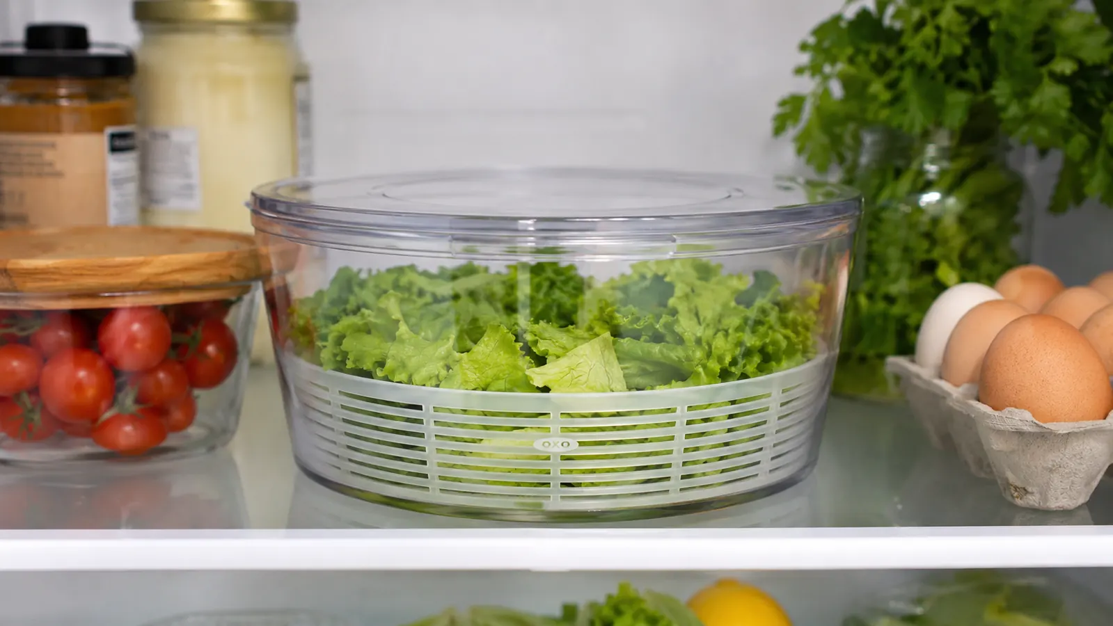 An open refrigerator shelf showing a salad spinner bowl covered with its lid storing crisp greens inside