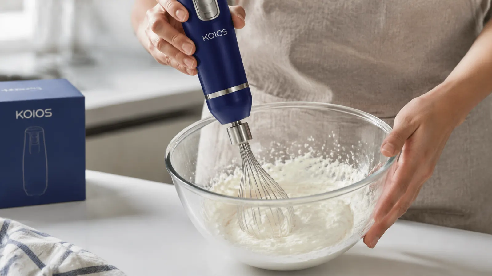 Close-up of the KOIOS whisk attachment sitting in a glass bowl of partially whipped cream, showing the wire whisk and motor connection point