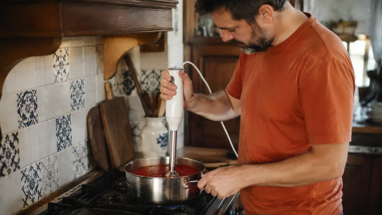 Home cook standing at the stove blending a thick red tomato sauce directly in a stainless saucepan, casual kitchen setting