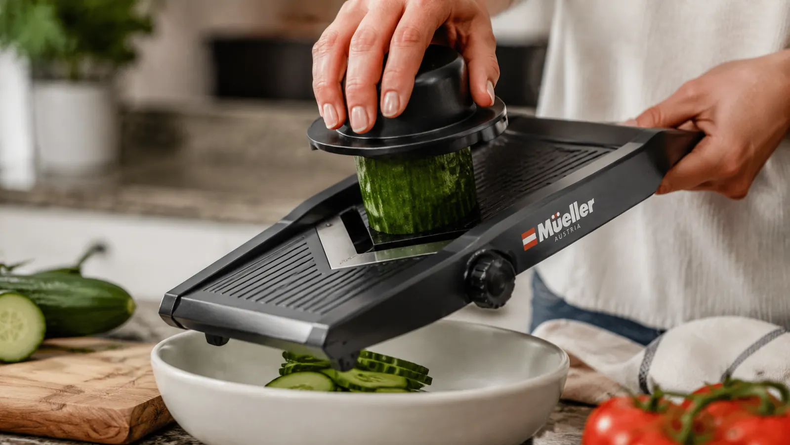 Hand guiding a cucumber through the Mueller mandoline slicer using the included food holder, producing uniform slices into a bowl below