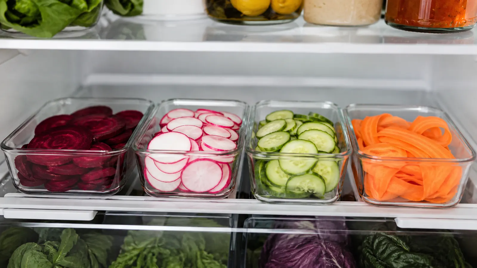 Glass meal-prep containers filled with thinly sliced vegetables including beets, radishes, and cucumbers arranged in a refrigerator