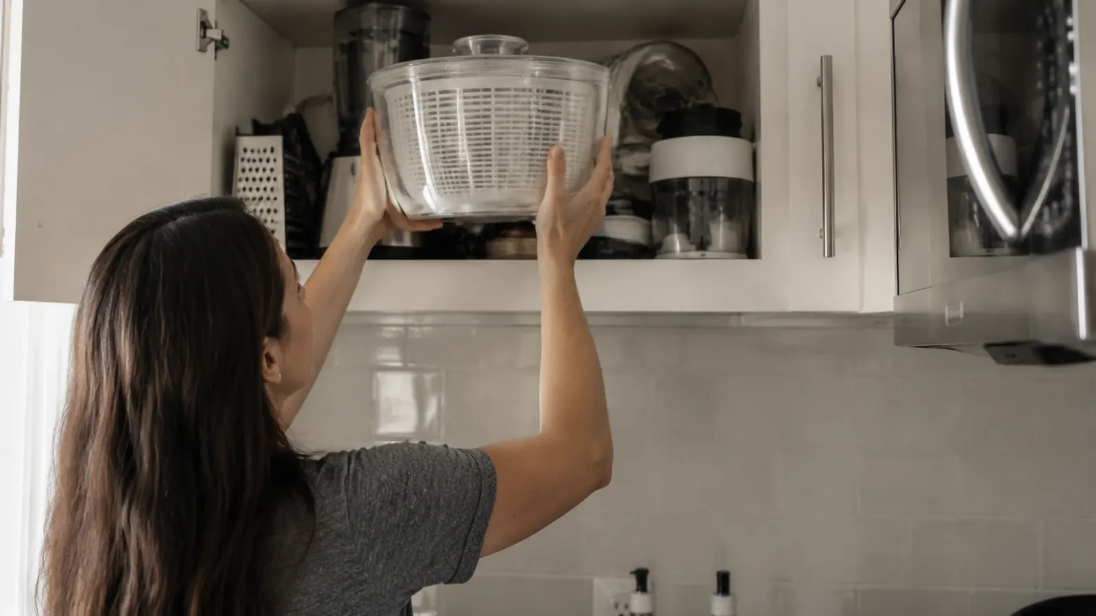 Person storing a large salad spinner in an overhead kitchen cabinet alongside other mixing bowls