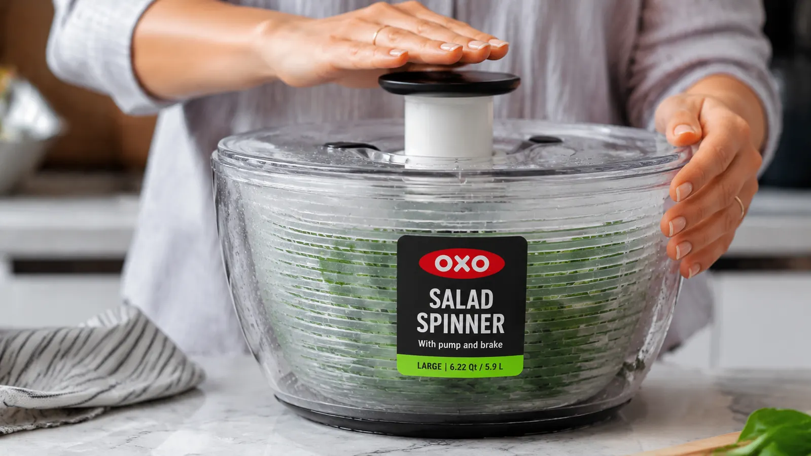Hand pressing down on the OXO salad spinner pump button with water droplets visible inside the clear bowl